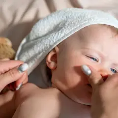 Delikatna pielęgnacja noworodka: mama delikatnie dotyka policzka dziecka w białej sapce, obok leży szczotka do włosów.