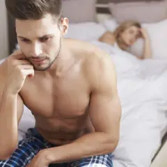 Mężczyzna w łóżku zamyślony, zmartwiony. Kobieta w tle. Problemy z libido u mężczyzn?
