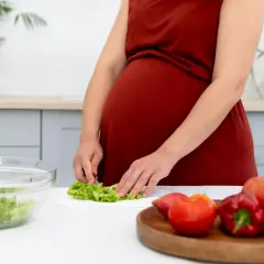 Ciężarna kobieta przygotowuje sałatkę, dbając o to, czego nie można jeść w ciąży. Na blacie leżą świeże warzywa.