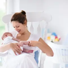 Uśmiechnięta mama kołysze noworodka. W tle widać łóżeczko i kwiaty. To obrazek dla tych, którzy szukają informacji, jak zatrzymać laktację.