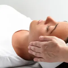 Relaks podczas zabiegu reiki. Delikatne dłonie terapeuty przykładane do głowy kobiety, która leży z zamkniętymi oczami.