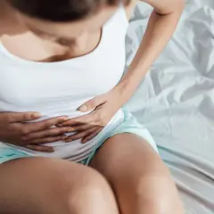 Kobieta w białej koszulce i turkusowych spodenkach trzyma się za brzuch, odczuwając ból podbrzusza w ciąży.