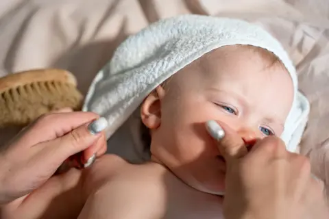 Delikatna pielęgnacja noworodka: mama delikatnie dotyka policzka dziecka w białej sapce, obok leży szczotka do włosów.