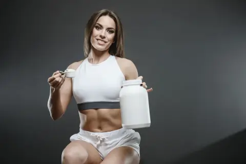 Uśmiechnięta sportsmenka z brzuchem jak z żelazka trzyma słoik i miarkę z proszkiem. Kreatyna co daje? Siłę i energię do treningów!
