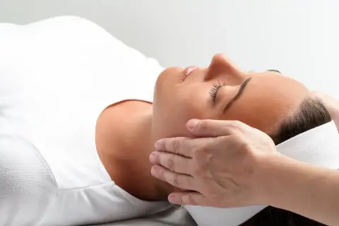 Relaks podczas zabiegu reiki. Delikatne dłonie terapeuty przykładane do głowy kobiety, która leży z zamkniętymi oczami.