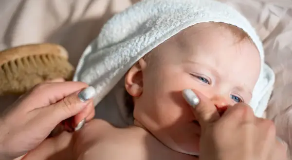 Delikatna pielęgnacja noworodka: mama delikatnie dotyka policzka dziecka w białej sapce, obok leży szczotka do włosów.