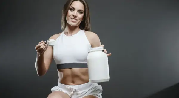 Uśmiechnięta sportsmenka z brzuchem jak z żelazka trzyma słoik i miarkę z proszkiem. Kreatyna co daje? Siłę i energię do treningów!