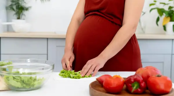 Ciężarna kobieta przygotowuje sałatkę, dbając o to, czego nie można jeść w ciąży. Na blacie leżą świeże warzywa.
