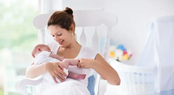 Uśmiechnięta mama kołysze noworodka. W tle widać łóżeczko i kwiaty. To obrazek dla tych, którzy szukają informacji, jak zatrzymać laktację.