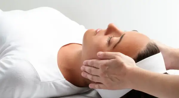 Relaks podczas zabiegu reiki. Delikatne dłonie terapeuty przykładane do głowy kobiety, która leży z zamkniętymi oczami.