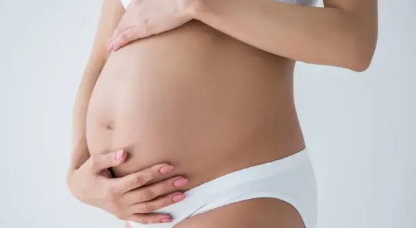 Kobieta w białej bieliźnie obejmuje dłońmi swój twardy brzuch w ciąży.