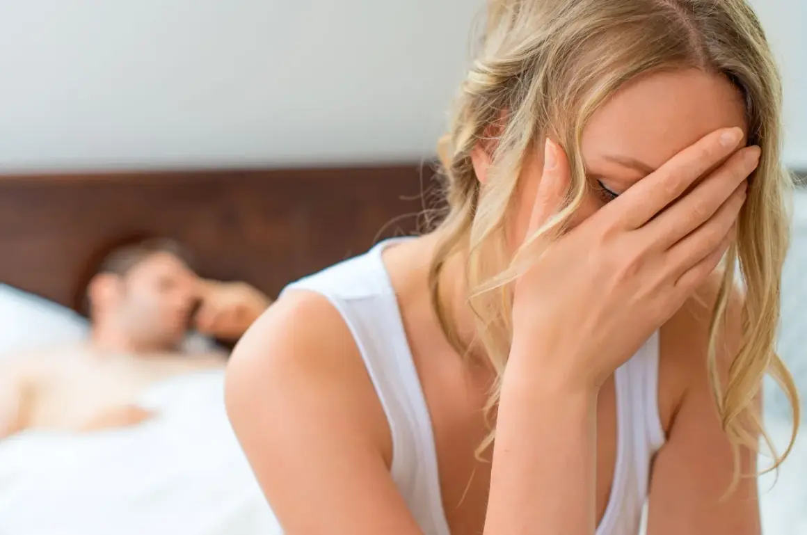 Kobieta w bieliźnie z ręką na twarzy, wyrażająca ból podczas stosunku przy wejściu do pochwy. W tle niewyraźny mężczyzna w łóżku.