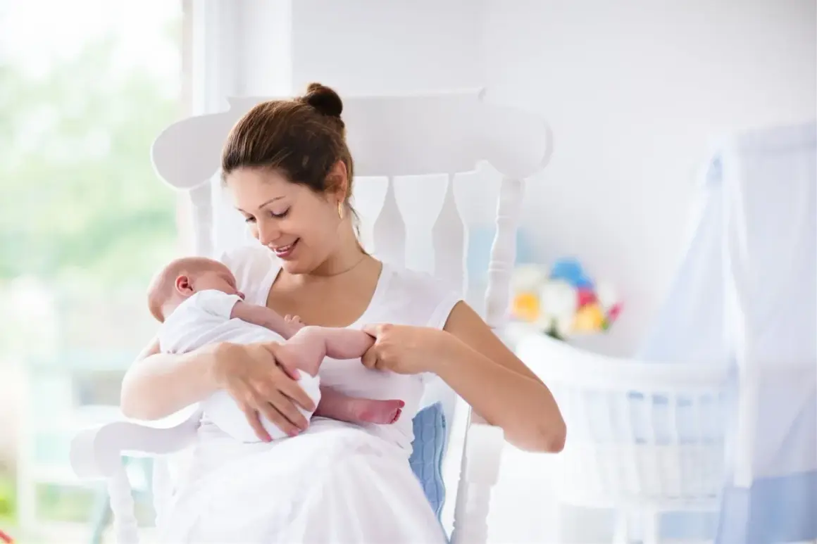Uśmiechnięta mama kołysze noworodka. W tle widać łóżeczko i kwiaty. To obrazek dla tych, którzy szukają informacji, jak zatrzymać laktację.