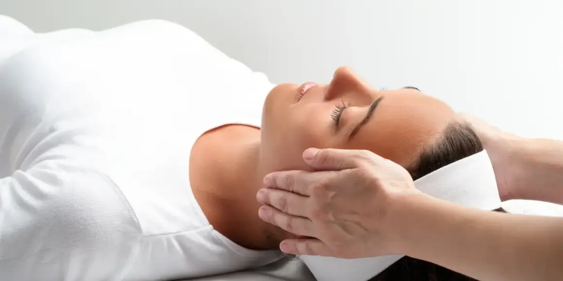Relaks podczas zabiegu reiki. Delikatne dłonie terapeuty przykładane do głowy kobiety, która leży z zamkniętymi oczami.