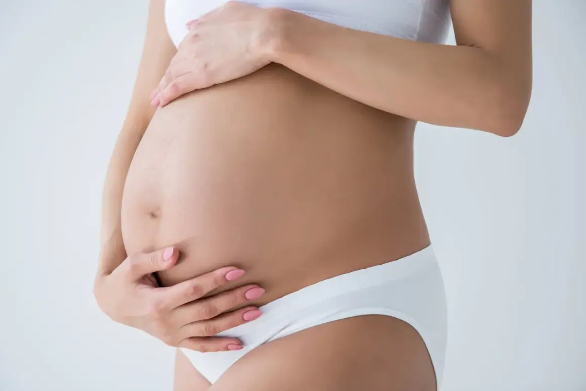 Kobieta w białej bieliźnie obejmuje dłońmi swój twardy brzuch w ciąży.