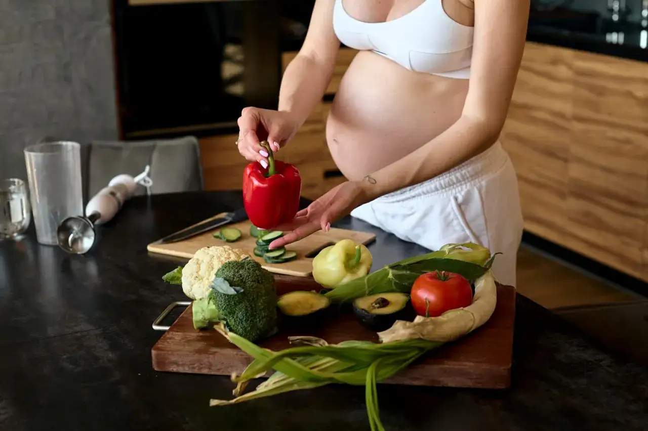 Ciężarna kobieta przygotowuje zdrowe jedzenie, ale pamiętaj, czego nie można jeść w ciąży. Na stole warzywa: papryka, brokuł, kalafior, awokado, pomidor.
