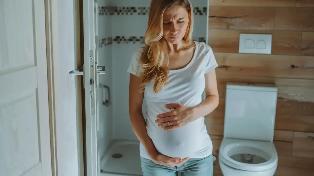 Kobieta w ciąży trzyma się za brzuch, odczuwając ból podbrzusza. W tle łazienka z toaletą.