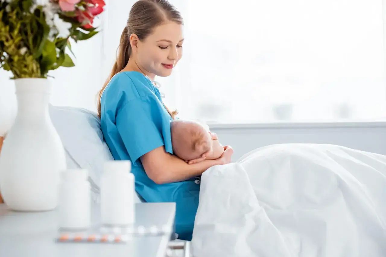 Kobieta w niebieskim fartuchu karmi piersią dziecko. Na stoliku obok leki, w tym ibuprofen.