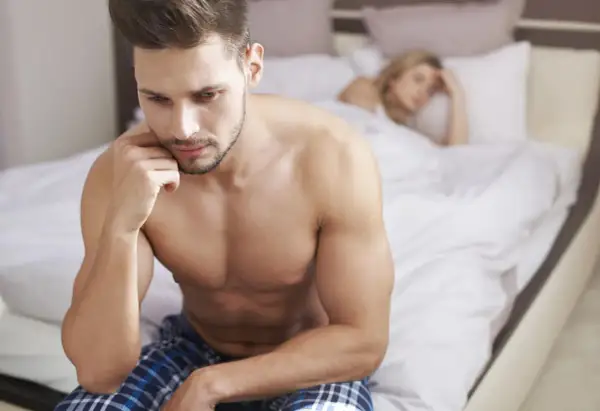 Mężczyzna w łóżku zamyślony, zmartwiony. Kobieta w tle. Problemy z libido u mężczyzn?