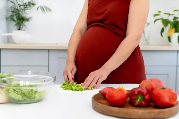 Ciężarna kobieta przygotowuje sałatkę, dbając o to, czego nie można jeść w ciąży. Na blacie leżą świeże warzywa.