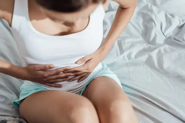 Kobieta w białej koszulce i turkusowych spodenkach trzyma się za brzuch, odczuwając ból podbrzusza w ciąży.